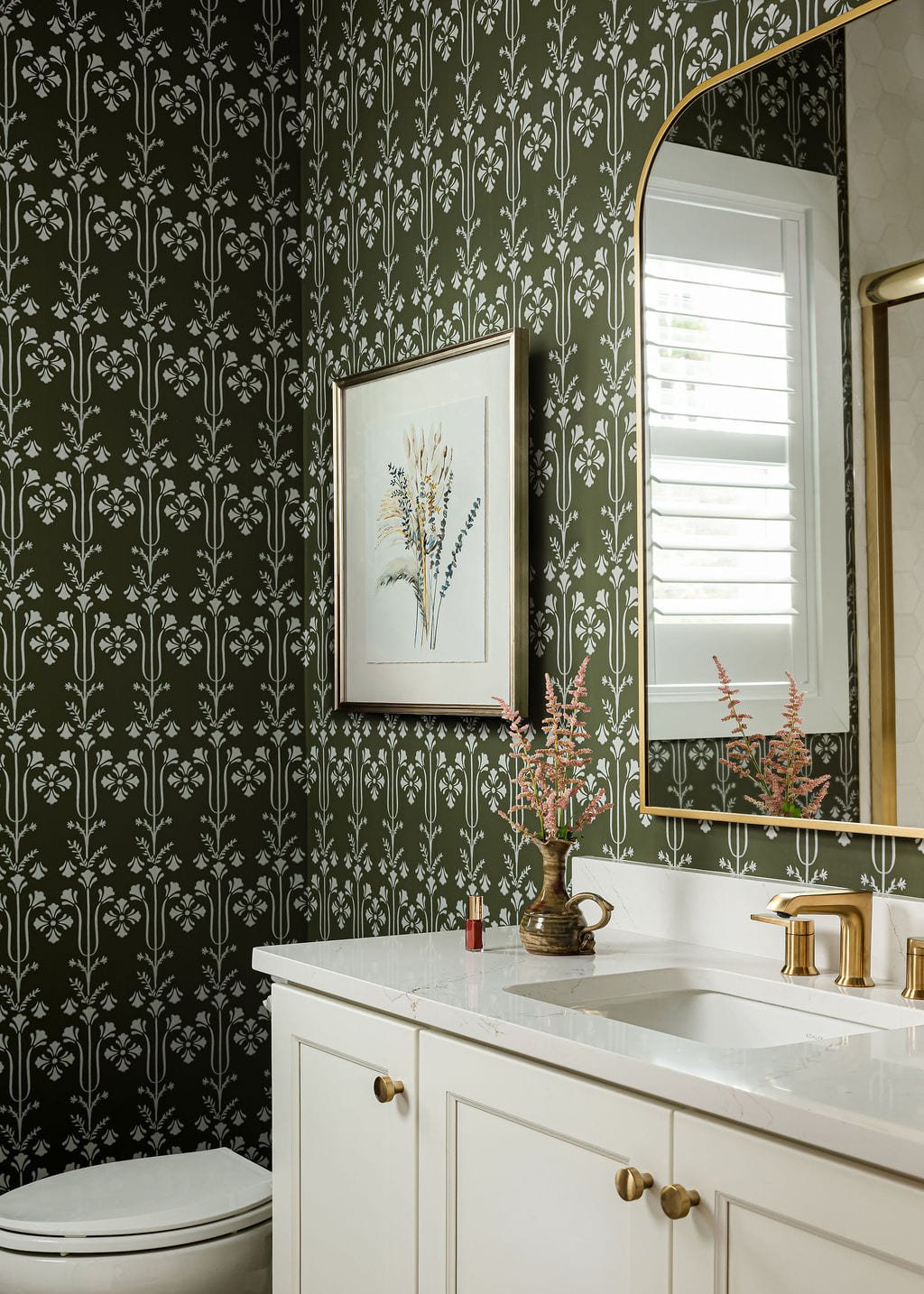 Powder room with dark botanical wallpaper and a brass arched mirror