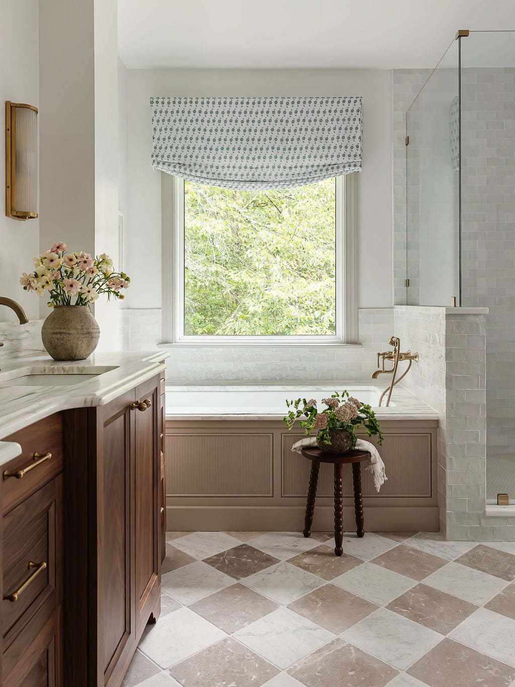 Soaking tub beneath a patterned roman shade with checkered stone flooring