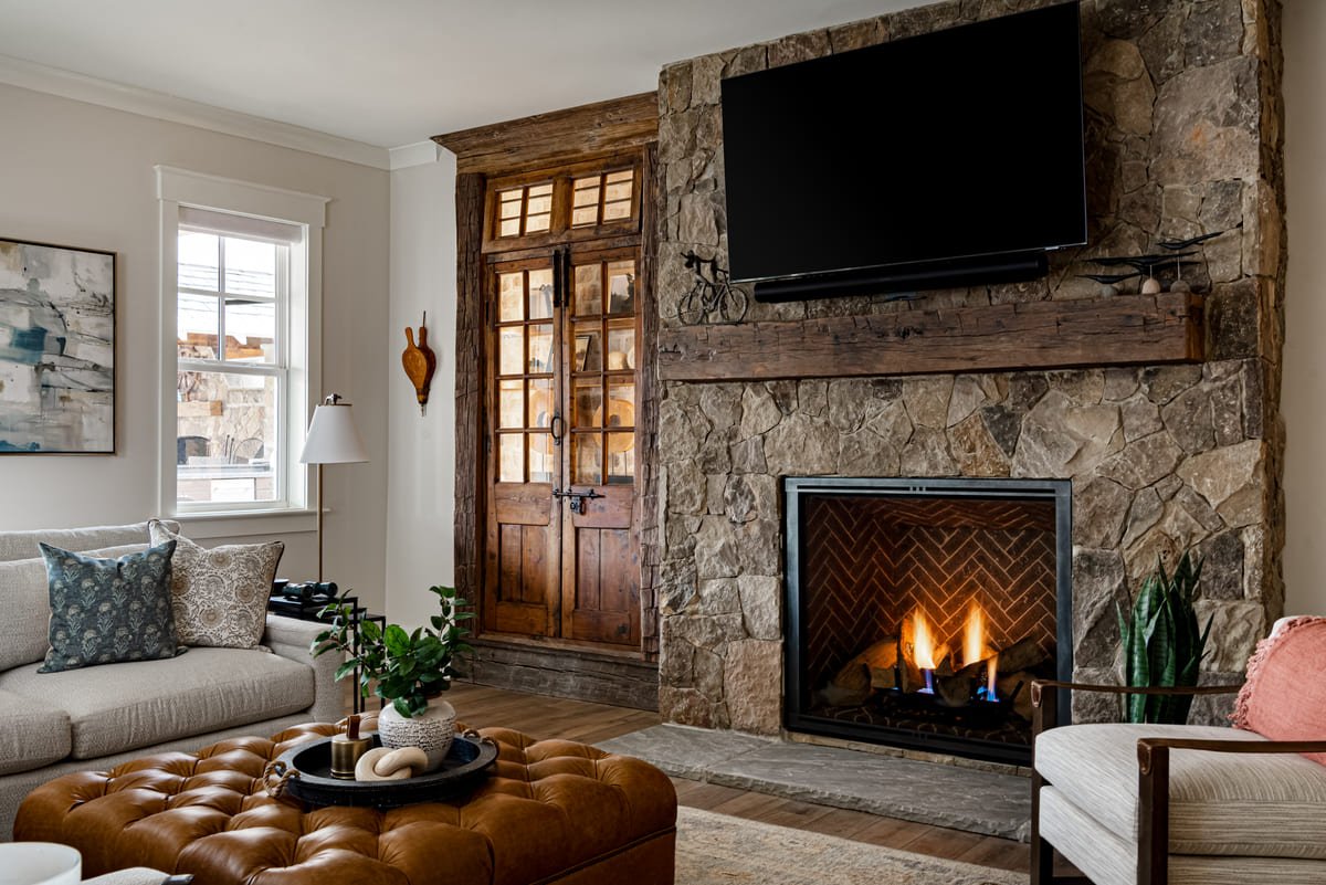 Stone fireplace lit beside a tufted leather ottoman in a lake house living room